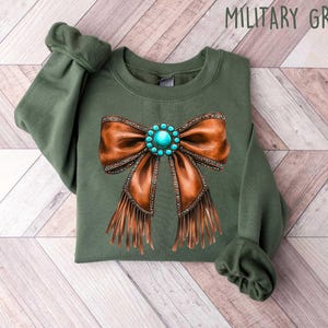 May include: A green sweatshirt with a brown leather bow with turquoise gemstone embellishments and fringe. The sweatshirt is labeled "Military Green".