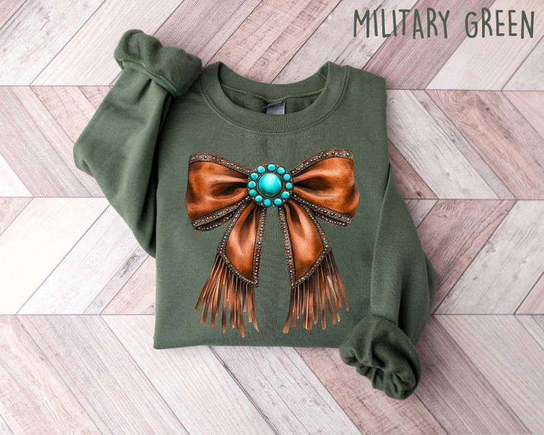May include: A green sweatshirt with a brown leather bow with turquoise gemstone embellishments and fringe. The sweatshirt is labeled "Military Green".