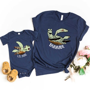 May include: Matching navy blue shirts featuring a cartoon turtle design. The adult shirt says "DUDE" and the baby shirt says "LIL DUDE".