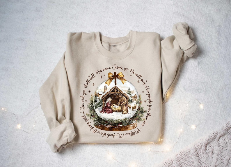 May include: Beige sweatshirt featuring a Christmas-themed graphic. The graphic is a snow globe with a nativity scene inside, surrounded by text from Matthew 1:21. The design includes a gold bow at the top of the snow globe.
