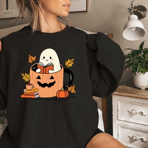May include: Black sweatshirt featuring a graphic of a ghost reading a book inside a pumpkin mug. The ghost is white with a smiling face and the pumpkin mug is orange with a black jack-o-lantern face. There are autumn leaves and books around the mug.