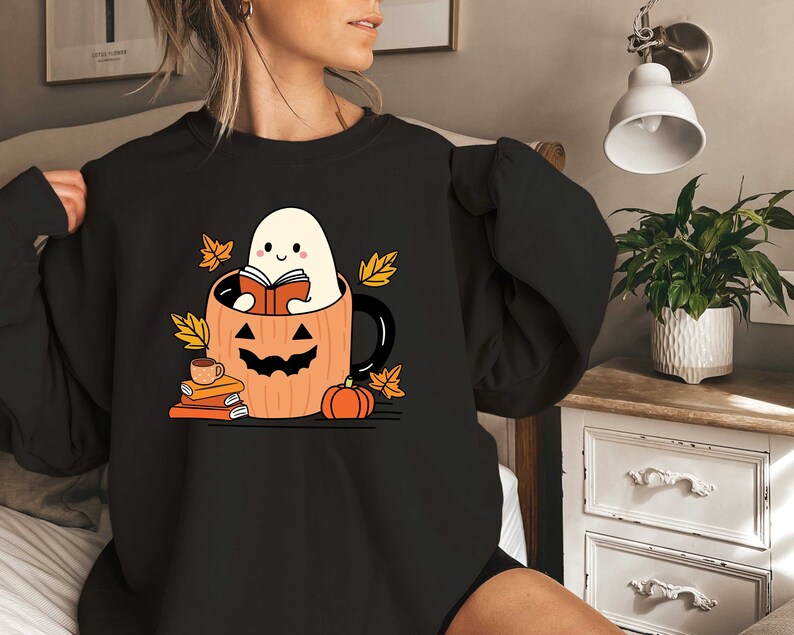 May include: Black sweatshirt featuring a graphic of a ghost reading a book inside a pumpkin mug. The ghost is white with a smiling face and the pumpkin mug is orange with a black jack-o-lantern face. There are autumn leaves and books around the mug.