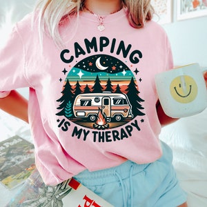 May include: Pink t-shirt with a retro camping design. The design features a camper van, a campfire, trees, stars, and a crescent moon. The text on the shirt reads "Camping is my therapy."