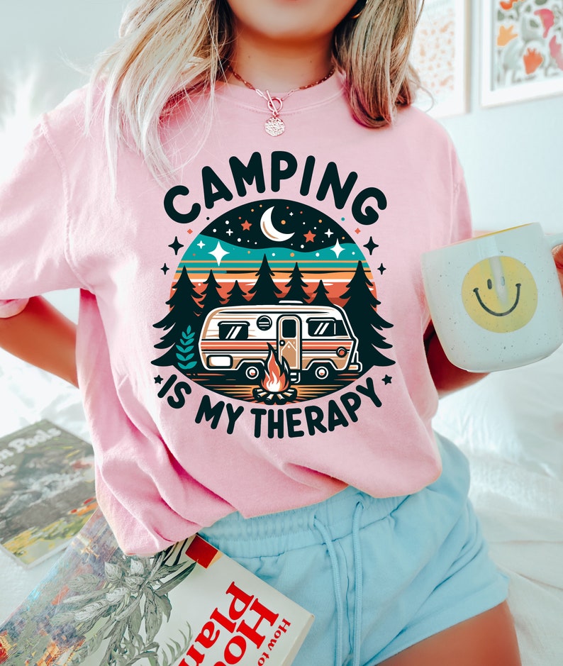 May include: Pink t-shirt with a retro camping design. The design features a camper van, a campfire, trees, stars, and a crescent moon. The text on the shirt reads "Camping is my therapy."