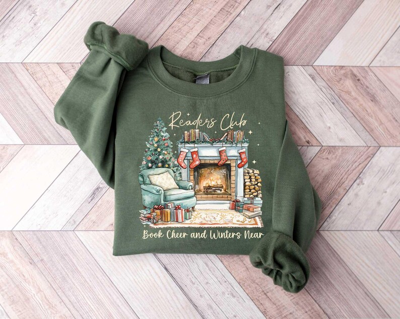 K&ouml;nnte beinhalten: Ein gr&uuml;nes Sweatshirt mit einem weihnachtlichen Design, das einen Kamin, einen Weihnachtsbaum und B&uuml;cher zeigt. Der Text "Readers Club" befindet sich &uuml;ber dem Kamin und "Book Cheer and Winters Near" unter dem Kamin.