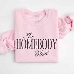 May include: A pale pink sweatshirt featuring the words "The Homebody Club" in black script. The long sleeves are folded and extend outwards. The sweatshirt is made of a soft fabric.