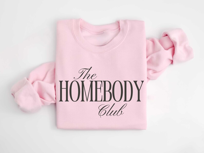 May include: A pale pink sweatshirt featuring the words "The Homebody Club" in black script. The long sleeves are folded and extend outwards. The sweatshirt is made of a soft fabric.
