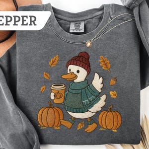 May include: Pepper-colored sweatshirt featuring an embroidered duck wearing a hat, scarf, and boots, holding a coffee cup, and walking past pumpkins and fall leaves. The word "PEPPER" is in a white rectangle in the upper left corner.