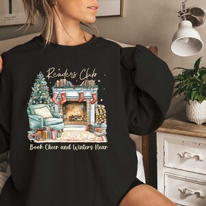K&ouml;nnte beinhalten: Schwarzes Sweatshirt mit einem Grafikdruck eines Kamins, B&uuml;chern und eines Weihnachtsbaums. Der Text "Readers Club" befindet sich &uuml;ber dem Grafikdruck und "Book Cheer and Winters Near" darunter.