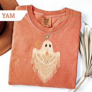 May include: A coral-colored Comfort Colors t-shirt featuring a ghost graphic with a floral design and a crescent moon. The shirt has a round neckline and short sleeves. A gold necklace with a pendant is draped on the shirt.