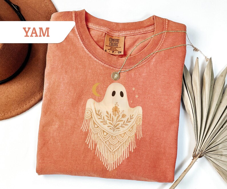 May include: A coral-colored Comfort Colors t-shirt featuring a ghost graphic with a floral design and a crescent moon. The shirt has a round neckline and short sleeves. A gold necklace with a pendant is draped on the shirt.