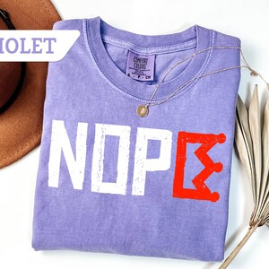 May include: A violet Comfort Colors t-shirt with the word "NOPE" in white block letters and a red crown graphic. A gold necklace with a pendant is draped across the shirt. A brown hat and dried plant are in the background.