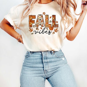 Może przedstawiać: Kremowa koszulka z napisem "Fall vibes" w brązowych i pomarańczowych literach. Litery są ozdobione jesiennymi motywami, takimi jak liście, żołędzie i dynie.