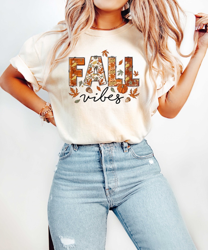 Może przedstawiać: Kremowa koszulka z napisem "Fall vibes" w brązowych i pomarańczowych literach. Litery są ozdobione jesiennymi motywami, takimi jak liście, żołędzie i dynie.