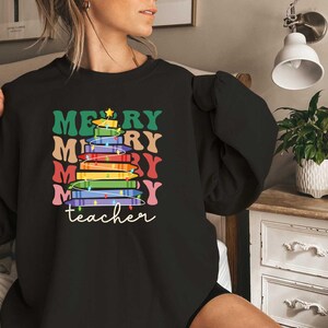 May include: Black sweatshirt with a colorful Christmas tree design made of books. The words "MERRY" and "teacher" are printed on the front in various colors. A star tops the tree.