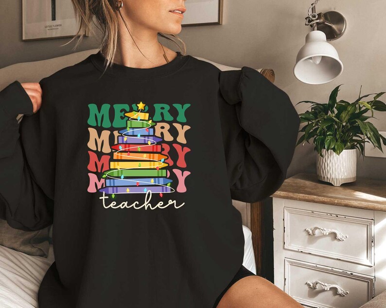 May include: Black sweatshirt with a colorful Christmas tree design made of books. The words "MERRY" and "teacher" are printed on the front in various colors. A star tops the tree.