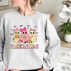 May include: A gray sweatshirt with a graphic of three cartoon ghosts wearing pink bows and holding various items. The text "PANTASMAS" is printed below the ghosts.