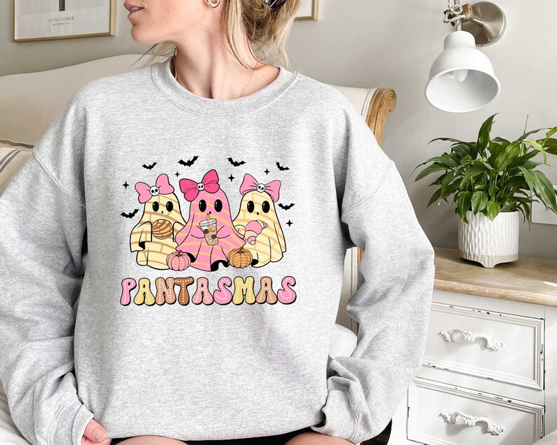 May include: A gray sweatshirt with a graphic of three cartoon ghosts wearing pink bows and holding various items. The text "PANTASMAS" is printed below the ghosts.