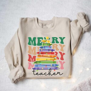 May include: Beige sweatshirt with a colorful Christmas tree design made of crayons. The words "MERRY" and "teacher" are printed on the front in various colors and fonts. A festive and fun design for teachers.