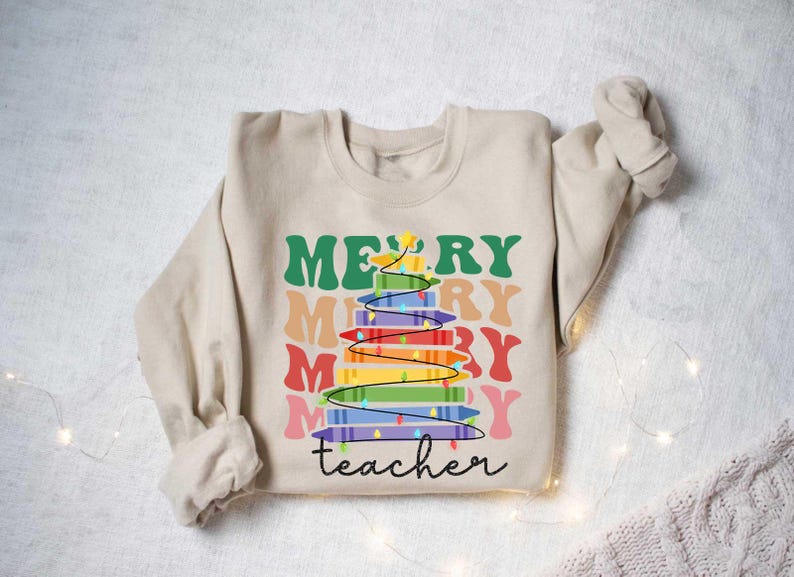 May include: Beige sweatshirt with a colorful Christmas tree design made of crayons. The words "MERRY" and "teacher" are printed on the front in various colors and fonts. A festive and fun design for teachers.