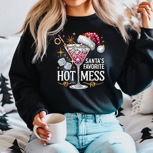 May include: A black sweatshirt with a festive design featuring a martini glass with a red liquid, ice cubes, and a Santa hat. The text "Santa's Favorite Hot Mess" is printed in gold lettering.