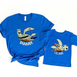 May include: Two blue t-shirts featuring a sea turtle design. The larger shirt reads "Dude" and the smaller shirt reads "Lil Dude".