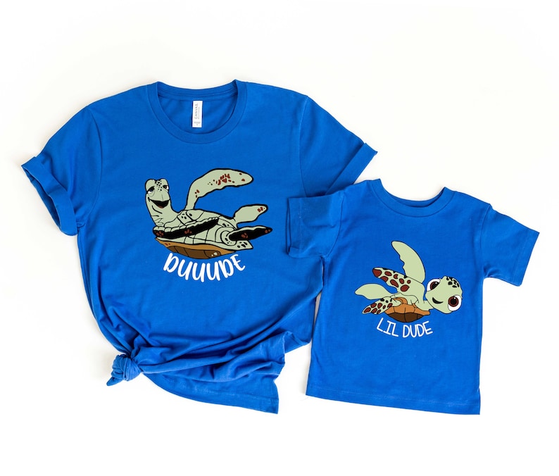 May include: Two blue t-shirts featuring a sea turtle design. The larger shirt reads "Dude" and the smaller shirt reads "Lil Dude".