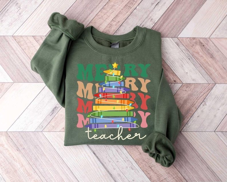 May include: A dark green sweatshirt with the words "MERRY teacher" in a colorful, stacked design resembling a Christmas tree made of books. The design includes a star and string lights.