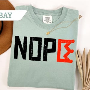 May include: A light green t-shirt with the word "NOPE" in distressed black letters and a red Greek letter. A gold necklace and a brown hat are also visible.