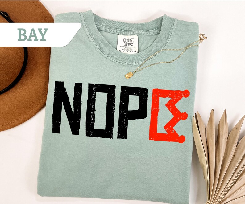 May include: A light green t-shirt with the word "NOPE" in distressed black letters and a red Greek letter. A gold necklace and a brown hat are also visible.