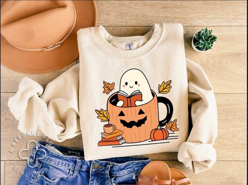 May include: A cream-coloured sweatshirt featuring a cute ghost reading a book inside a pumpkin mug. The ghost is smiling and holding a book. The mug is surrounded by autumn leaves and a stack of books.