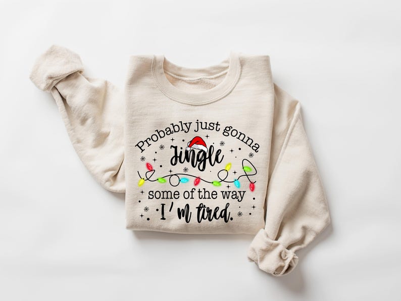 Peut inclure: Sweat-shirt beige avec l'inscription "Probably just gonna jingle some of the way. I'm tired." Le mot "Jingle" est surmont&eacute; d'un bonnet de P&egrave;re No&euml;l. Une guirlande de lumi&egrave;res de No&euml;l color&eacute;es est en dessous.