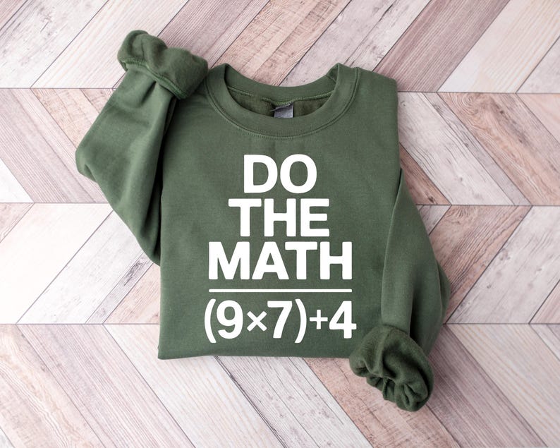 May include: An olive green sweatshirt with the text "DO THE MATH" in bold white letters. Below the text is the math equation (9x7)+4. The sweatshirt is folded, with the sleeves cuffed.