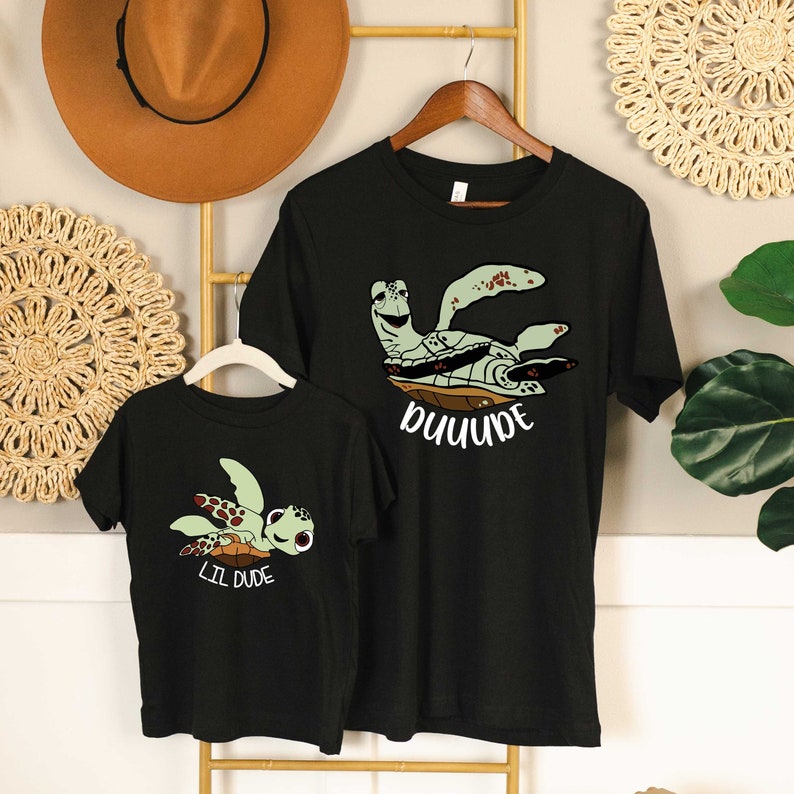 May include: Two black t-shirts featuring a green sea turtle graphic. The larger shirt reads "DUDE" and the smaller shirt reads "LIL DUDE".