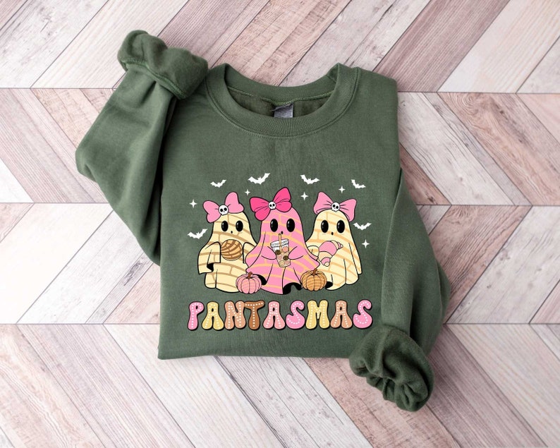 May include: Green sweatshirt with a graphic of three cartoon ghosts wearing pink bows and holding drinks and a pumpkin. The text "PANTASMAS" is printed in a retro font below the ghosts.