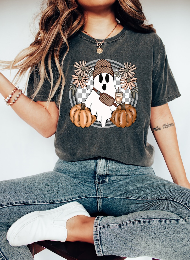 May include: A dark gray t-shirt with a white ghost wearing a brown and white beanie, a brown and white crossbody bag, and holding a brown and white cup. The ghost is surrounded by brown pumpkins and pink flowers. The background is a checkered pattern.