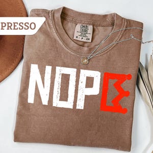 May include: Espresso-colored t-shirt with the word "NOPE" in white, distressed font. A red graphic of a crown-like symbol is on the right. A silver necklace with a small pendant is on the shirt.