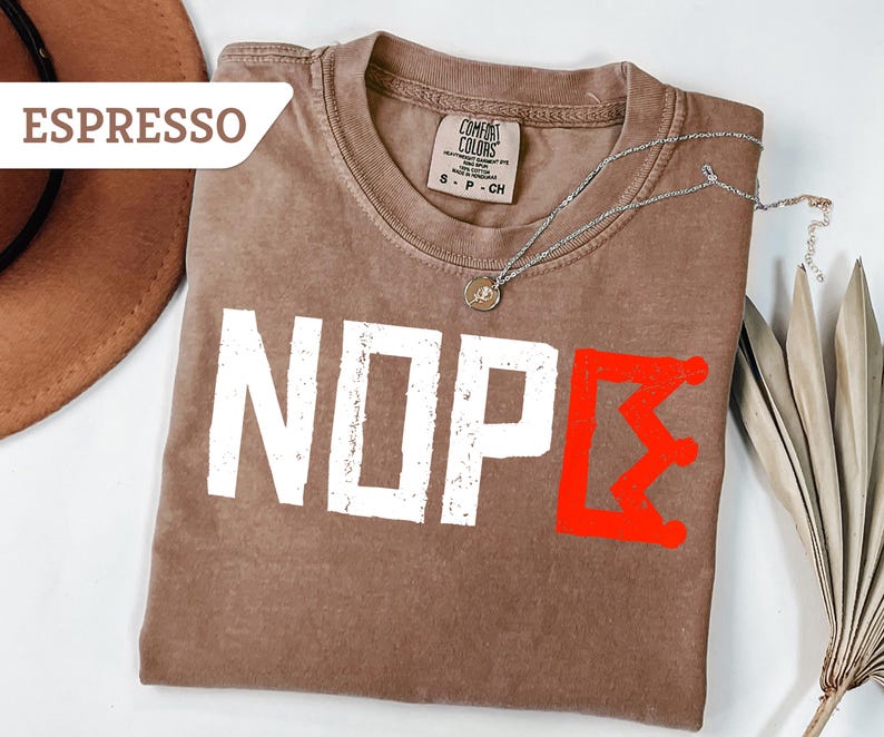 May include: Espresso-colored t-shirt with the word "NOPE" in white, distressed font. A red graphic of a crown-like symbol is on the right. A silver necklace with a small pendant is on the shirt.