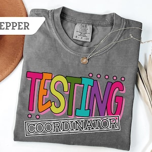 May include: A gray t-shirt with the words "TESTING COORDINATOR" in colorful block letters. The shirt has a crew neck and short sleeves. A gold necklace with a pendant is draped across the shirt. A brown hat is visible in the background.
