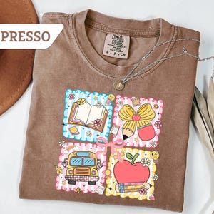 May include: Espresso-colored t-shirt with a colorful graphic featuring a book, pencil, school bus, and apple. The design is framed by a decorative border with flowers and stars. A silver necklace is draped across the shirt.