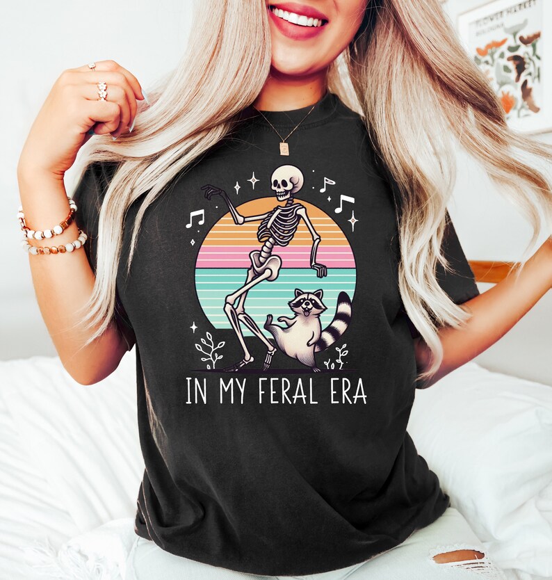 以下が含まれることがあります： レトロなスタイルのグラフィックがプリントされた黒いTシャツ。グラフィックは、星と音符が描かれた夕焼けの背景に、スケルトンがアライグマと踊っている様子が描かれています。グラフィックの下には「In My Feral Era」というテキストがプリントされています。