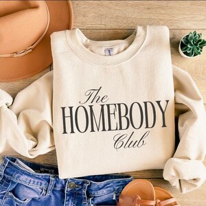 May include: A cream-coloured sweatshirt with the text "The Homebody Club" in black script. The sweatshirt is paired with blue denim shorts, a brown hat, and sandals. A small succulent plant is also visible.