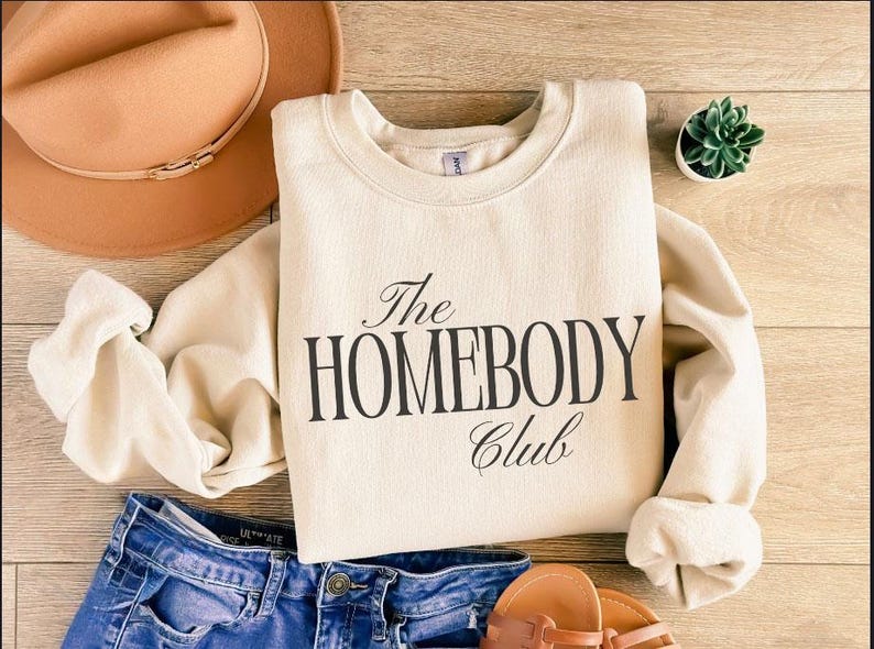 May include: A cream-coloured sweatshirt with the text "The Homebody Club" in black script. The sweatshirt is paired with blue denim shorts, a brown hat, and sandals. A small succulent plant is also visible.
