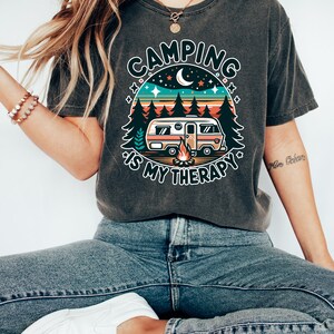 May include: A dark grey t-shirt with a retro-style graphic of a camper van in a forest setting. The graphic includes a campfire, stars, and a crescent moon. The text "Camping is my therapy" is printed below the graphic.