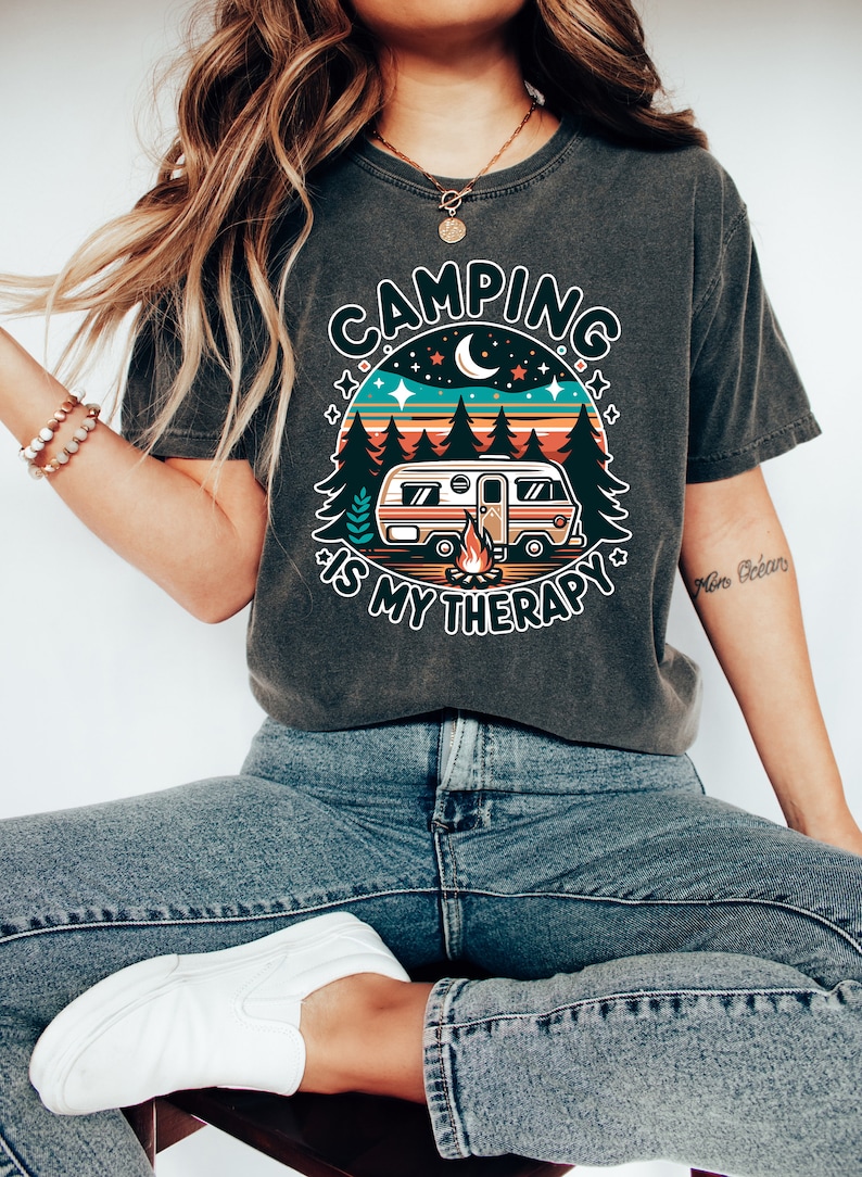 May include: A dark grey t-shirt with a retro-style graphic of a camper van in a forest setting. The graphic includes a campfire, stars, and a crescent moon. The text "Camping is my therapy" is printed below the graphic.