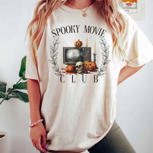 K&ouml;nnte beinhalten: Ein cremefarbenes T-Shirt mit einem Motiv aus einem Vintage-Fernseher, K&uuml;rbissen und einem Sch&auml;del. Der Text "Spooky Movie Club" ist auf das Shirt gedruckt.