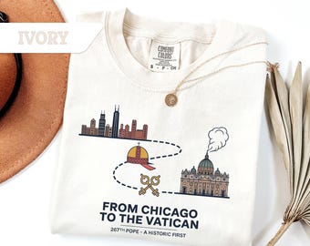 Könnte beinhalten: Elfenbeinfarbenes T-Shirt mit einem Grafikdesign, das die Skyline von Chicago, einen Papsthut, Schlüssel und den Vatikan zeigt. Der Text lautet "From Chicago to the Vatican" und "267th Pope - A Historic First". Eine goldene Halskette und ein brauner Hut sind ebenfalls sichtbar.