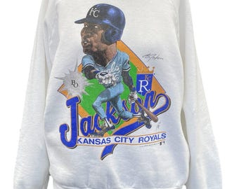 May include: White sweatshirt featuring a cartoon illustration of a baseball player in a blue helmet and uniform. The design includes the text "Jackson" and "Kansas City Royals" in blue and orange, with a baseball diamond graphic.