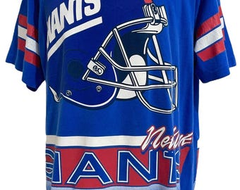 Könnte beinhalten: Blaues T-Shirt mit einem Football-Helm-Motiv. Das Shirt zeigt das Wort "GIANTS" in Weiß und Rot, wobei der Teamname wiederholt wird. Rote und weiße Streifen zieren die Ärmel. Ein Vintage-Sportartikel.