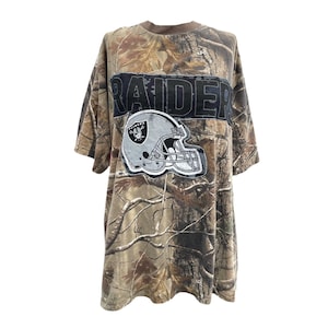 May include: A short-sleeve t-shirt featuring a brown and tan camouflage pattern. The front displays a large black rectangular patch with the word RAIDERS in bold letters, positioned above a graphic of a silver and black football helmet.